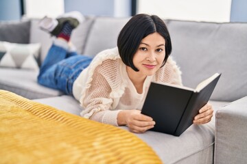 Young chinese woman reading book lying on sofa at home