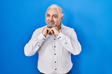 Middle age man with grey hair standing over blue background laughing nervous and excited with hands on chin looking to the side