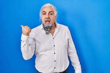 Middle age man with grey hair standing over blue background surprised pointing with hand finger to the side, open mouth amazed expression.