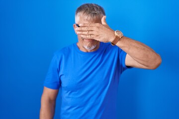 Hispanic man with grey hair standing over blue background smiling and laughing with hand on face covering eyes for surprise. blind concept.