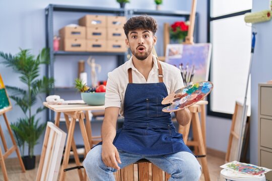 Arab man with beard painter sitting at art studio holding palette scared and amazed with open mouth for surprise, disbelief face