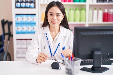 Young chinese woman pharmacist smiling confident holding glasses at pharmacy