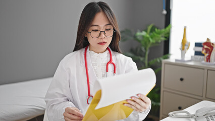 Young chinese woman doctor reading document on clipboard at clinic