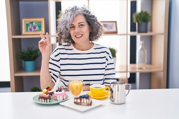 Middle age woman with grey hair eating pastries and drinking coffee for breakfast smiling with happy face winking at the camera doing victory sign. number two.