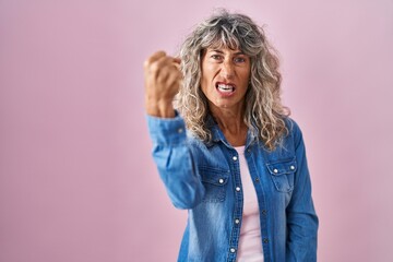 Middle age woman standing over pink background angry and mad raising fist frustrated and furious...