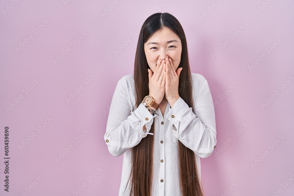 Poster chinese young woman standing over pink background laughing and embarrassed giggle covering mouth wit