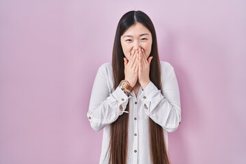 Chinese young woman standing over pink background laughing and embarrassed giggle covering mouth with hands, gossip and scandal concept