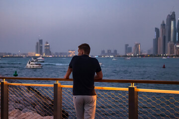 Rear view of young guy on embankment with view of skyscrapers Dubai UAE, looking around. Confident male tourist posing on urban district. Recreation and leisure activity concept. Copy ad text space