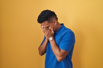 Young hispanic man standing over yellow background with sad expression covering face with hands...