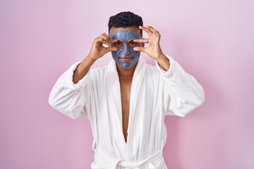 Young hispanic man wearing beauty face mask and bath robe trying to open eyes with fingers, sleepy and tired for morning fatigue