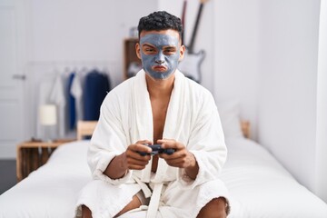 Young hispanic man wearing beauty face mask playing video games puffing cheeks with funny face. mouth inflated with air, catching air.