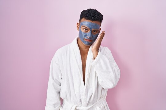 Young hispanic man wearing beauty face mask and bath robe thinking looking tired and bored with depression problems with crossed arms.