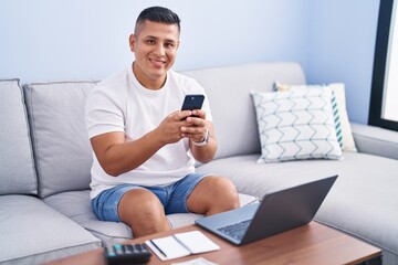 Young latin man using smartphone and laptop at home