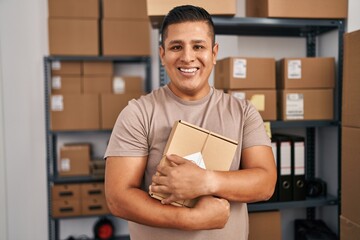 Young latin man ecommerce business worker hugging package at office
