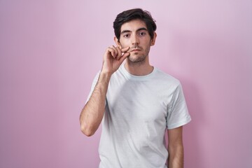 Young hispanic man standing over pink background mouth and lips shut as zip with fingers. secret...