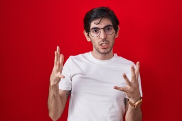 Naklejka premium Young hispanic man standing over red background crazy and mad shouting and yelling with aggressive expression and arms raised. frustration concept.