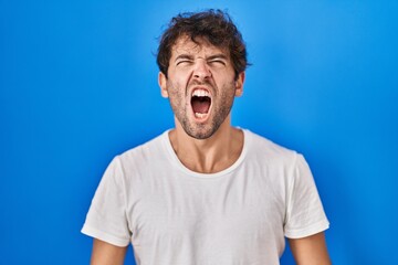 Hispanic young man standing over blue background angry and mad screaming frustrated and furious, shouting with anger. rage and aggressive concept.