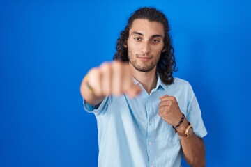 Young hispanic man standing over blue background punching fist to fight, aggressive and angry...