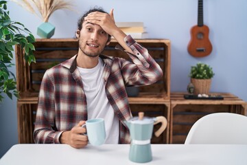 Young hispanic man drinking coffee from french coffee maker stressed and frustrated with hand on head, surprised and angry face