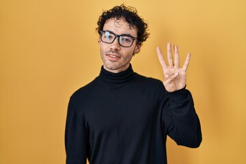 Hispanic man standing over yellow background showing and pointing up with fingers number four while smiling confident and happy.