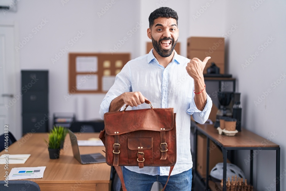 Wall mural Hispanic man with beard working at the office holding briefcase pointing thumb up to the side smiling happy with open mouth