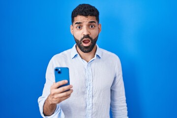Hispanic man with beard using smartphone typing message in shock face, looking skeptical and...