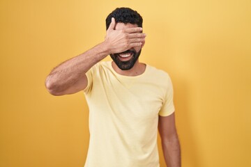 Hispanic man with beard standing over yellow background smiling and laughing with hand on face...