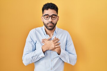 Hispanic man with beard standing over yellow background smiling with hands on chest with closed eyes and grateful gesture on face. health concept.