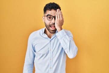 Hispanic man with beard standing over yellow background covering one eye with hand, confident smile...