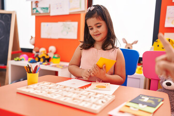 Adorable hispanic girl playing with vocabulary puzzle game sitting on table at kindergarten