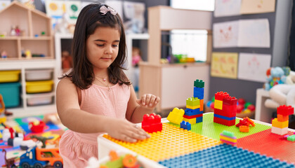 Adorable hispanic girl playing with construction blocks sitting on table at kindergarten
