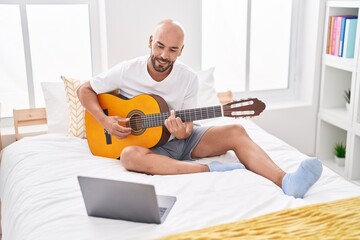 Young bald man having online classical guitar class sitting on bed at bedroom
