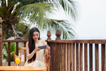 Fototapeta na wymiar Young woman with long hair sitting on terrace of beach hotel and watching videos on smartphone