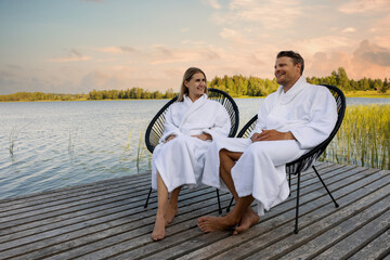 happy couple in white bathrobes relaxing on wooden lake footbridge after spa treatments at warm summer evening - obrazy, fototapety, plakaty