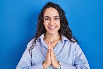 Young brunette woman standing over blue background praying with hands together asking for forgiveness smiling confident.