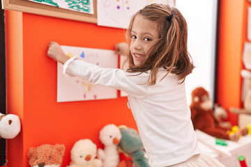 Adorable hispanic girl preschool student hanging draw on wall at kindergarten