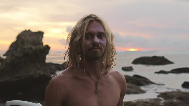 Portrait of attractive bearded blond Caucasian man looking into camera smiling on sea or ocean beach. Young male hipster millennial on beach at sunset. Long-haired blonde surfer hipster millennial