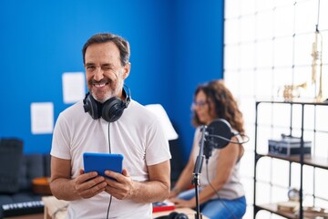 Middle age hispanic man at music studio using tablet winking looking at the camera with sexy expression, cheerful and happy face.