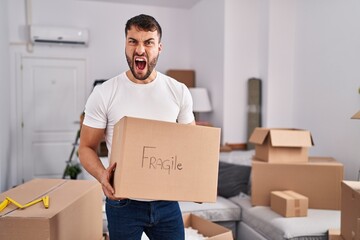Handsome hispanic man moving to a new home holding fragile box angry and mad screaming frustrated...