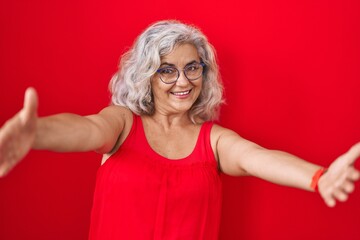 Middle age woman with grey hair standing over red background looking at the camera smiling with open arms for hug. cheerful expression embracing happiness.