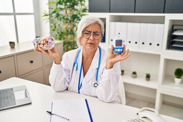 Middle age woman with grey hair wearing doctor uniform holding glucose monitor depressed and worry...
