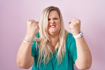 Caucasian plus size woman standing over pink background angry and mad raising fists frustrated and...