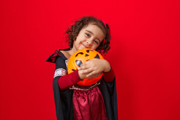 Adorable hispanic girl wearing halloween costume hugging pumpkin basket over isolated red background