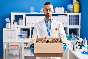 Young hispanic man working at scientist laboratory holding cardboard box relaxed with serious...