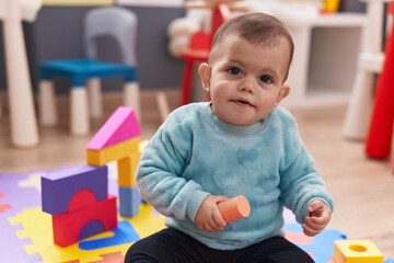 Adorable hispanic baby playing with construction blocks sitting on floor at kindergarten