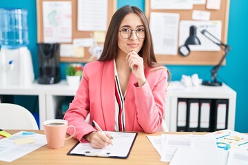 Young hispanic woman working at the office wearing glasses looking confident at the camera smiling with crossed arms and hand raised on chin. thinking positive.