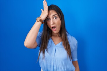 Young brunette woman standing over blue background surprised with hand on head for mistake, remember error. forgot, bad memory concept.