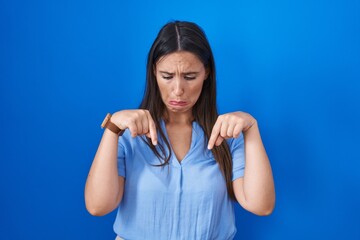 Young brunette woman standing over blue background pointing down looking sad and upset, indicating direction with fingers, unhappy and depressed.