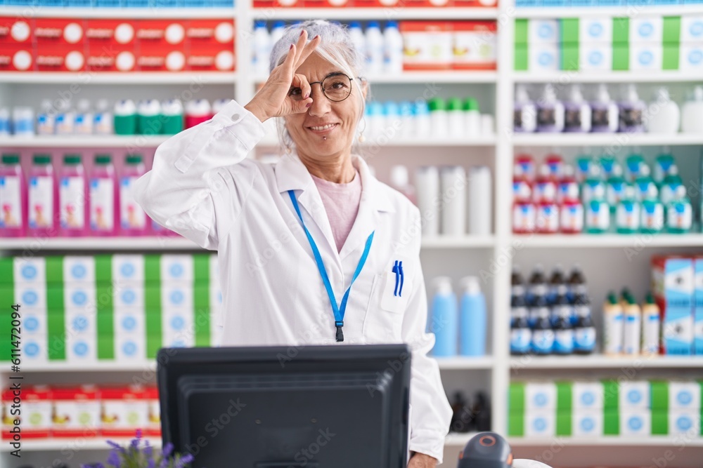 Poster Middle age woman with tattoos working at pharmacy drugstore doing ok gesture with hand smiling, eye looking through fingers with happy face.