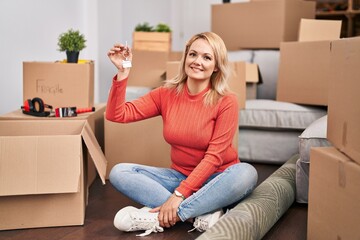 Blonde woman holding keys of new home sitting on the floor looking positive and happy standing and smiling with a confident smile showing teeth